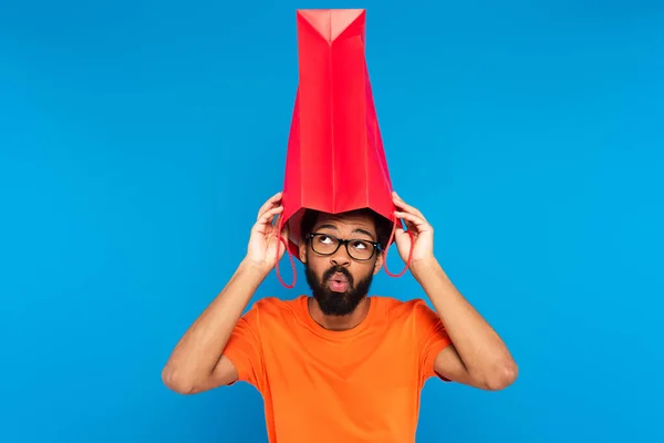 Surpris homme afro-américain dans des lunettes tenant sac à provisions au-dessus de la tête isolé sur bleu — Photo de stock