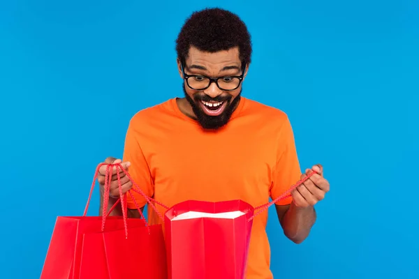 Excité homme afro-américain dans des lunettes regardant à l'intérieur du sac isolé sur bleu — Photo de stock