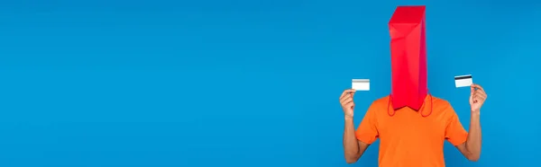 Hombre afroamericano con bolsa de compras en la cabeza celebración de tarjetas de crédito aisladas en azul, pancarta - foto de stock