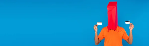 African american man with shopping bag on head holding credit cards isolated on blue, banner — Stock Photo