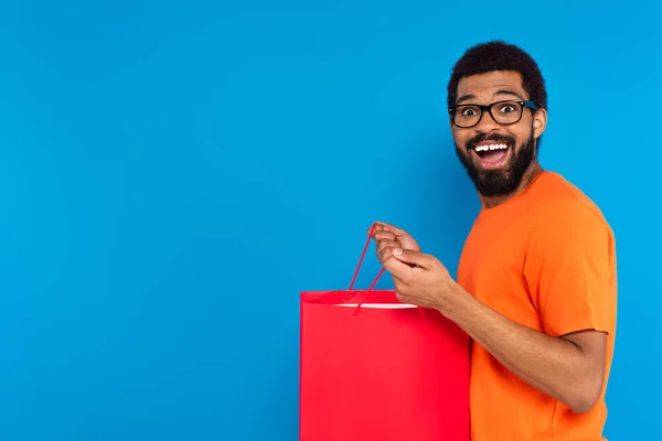 Animado afro-americano homem em óculos segurando saco de compras vermelho isolado em azul — Fotografia de Stock