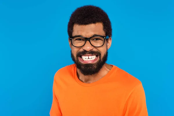 Homme afro-américain stressé dans des lunettes montrant des dents blanches isolées sur bleu — Photo de stock