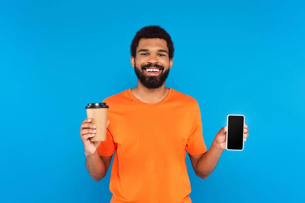 Allegro uomo afro americano in possesso di tazza di carta e smartphone isolato su blu — Foto stock