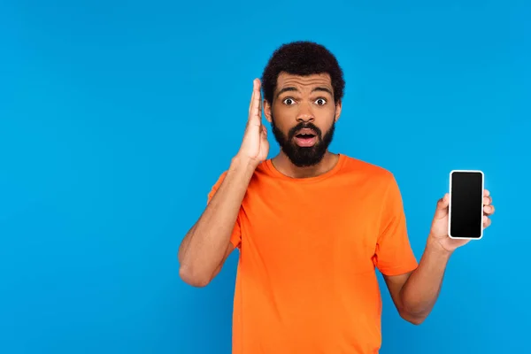 Sorprendido hombre afroamericano sosteniendo teléfono inteligente con pantalla en blanco aislado en azul - foto de stock