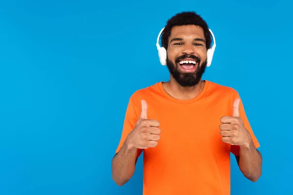 Alegre afroamericano hombre en auriculares inalámbricos escuchando música mientras muestra pulgares hacia arriba aislado en azul - foto de stock