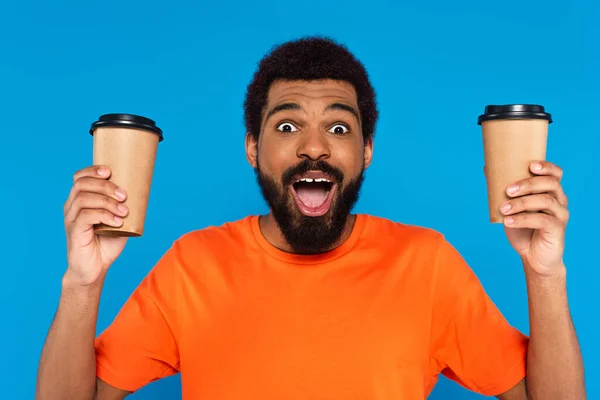 Étonné homme afro-américain tenant des tasses en papier isolé sur bleu — Photo de stock