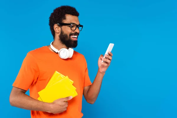 Homem americano africano alegre em fones de ouvido sem fio segurando livros e usando smartphone isolado em azul — Fotografia de Stock