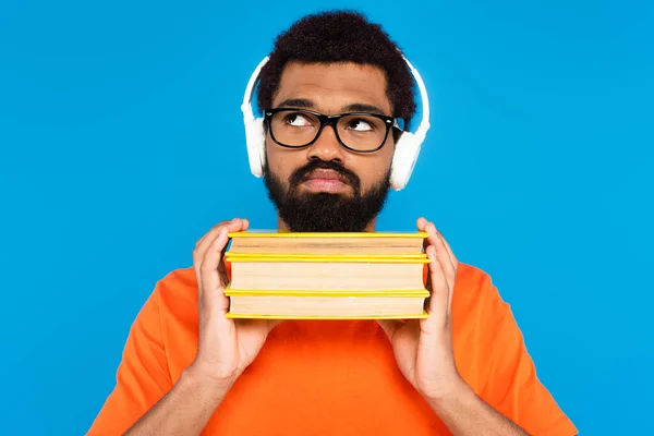 Uomo africano americano pensieroso in cuffie senza fili con libri isolati sul blu — Foto stock