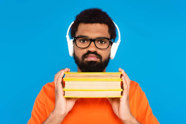 Bearded african american man in wireless headphones holding books isolated on blue — Stock Photo