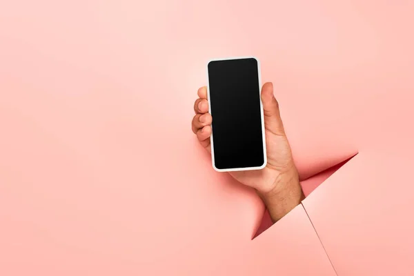 Partial view of african american man holding smartphone with blank screen on ripped pink background — Stock Photo
