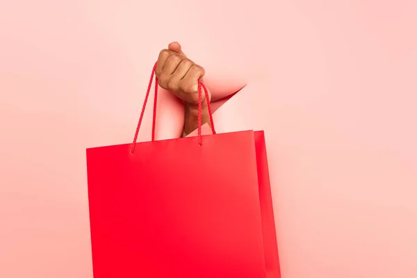 Vista cortada do homem afro-americano segurando saco de compras em fundo rosa rasgado — Fotografia de Stock