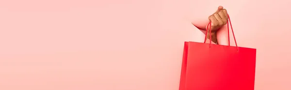 Partial view of african american man holding shopping bag on ripped pink background, banner — Stock Photo