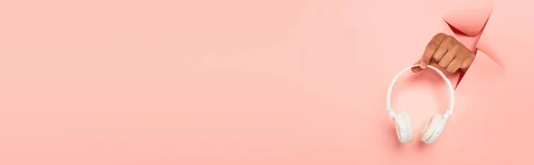 Vista parziale dell'uomo afroamericano che tiene cuffie wireless su sfondo rosa strappato, banner — Foto stock