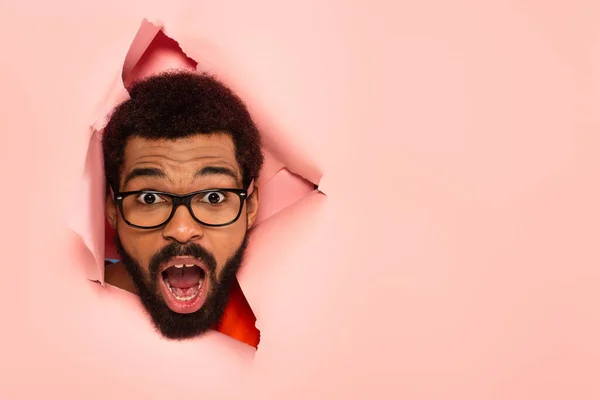 Shocked and bearded african american man with open mouth on ripped pink background — Stock Photo