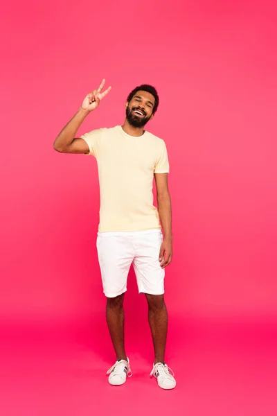 Comprimento total de homem americano africano alegre e barbudo mostrando sinal de paz em rosa — Fotografia de Stock