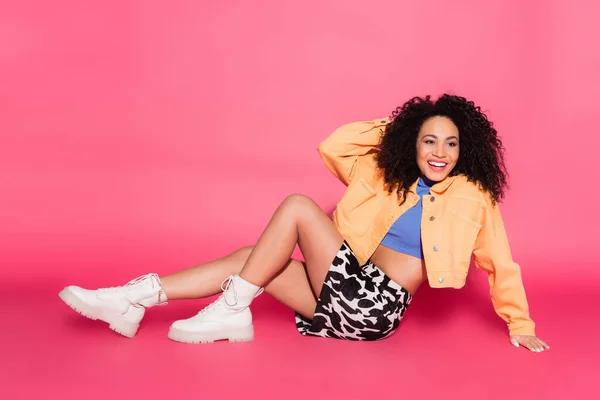 Full length of happy african american woman in crop top, jacket and skirt with animal print sitting on pink — Stock Photo