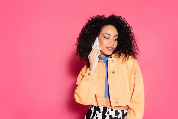 Curly african american woman in crop top and jacket talking on smartphone on pink — Stock Photo