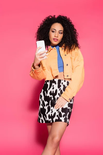 Lockige Afroamerikanerin in bauchfreiem Top und Jacke macht Selfie auf Smartphone auf rosa — Stockfoto