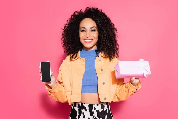 Curly african american woman in crop top and jacket holding smartphone with blank screen and present on pink — Stock Photo