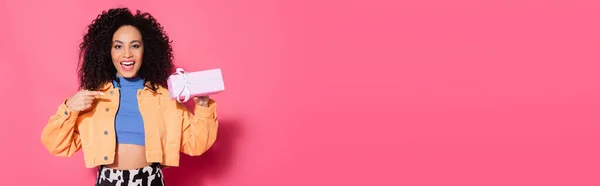 Amazed african american woman in crop top and jacket pointing at present on pink, banner — Stock Photo