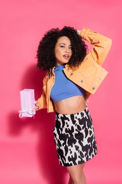 Curly african american woman in crop top, skirt and jacket holding present on pink — Stock Photo