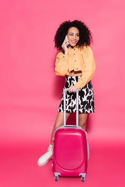 Full length of happy african american woman talking on smartphone near suitcase on pink — Stock Photo
