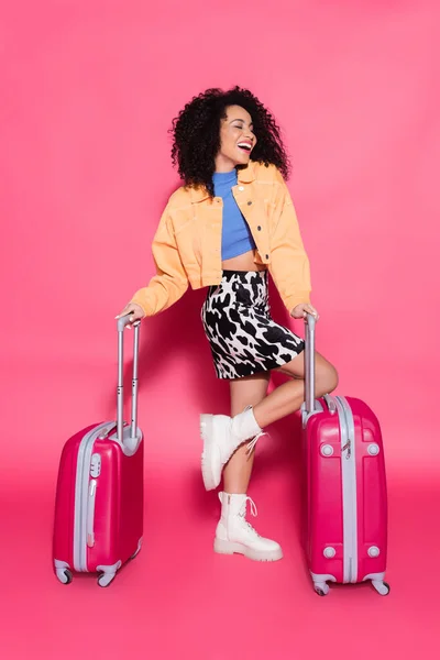 Full length of stylish african american woman smiling while posing with suitcases on pink — Stock Photo
