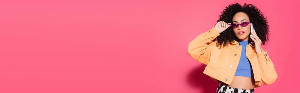 Curly african american woman in sunglasses talking on smartphone on pink, banner — Stock Photo