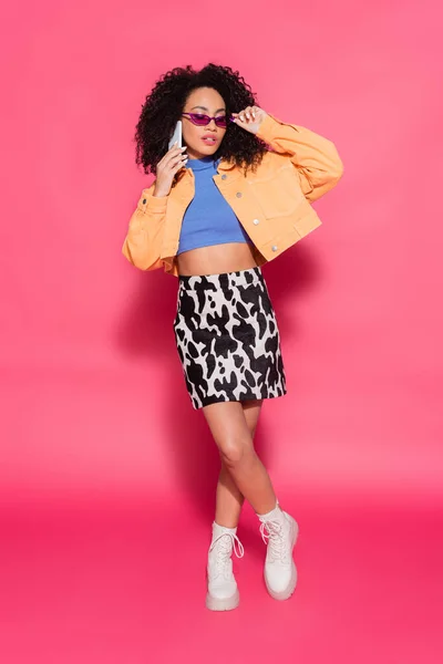 Повна довжина афроамериканської жінки, що налаштовує сонцезахисні окуляри і розмовляє на смартфоні на рожевому — стокове фото