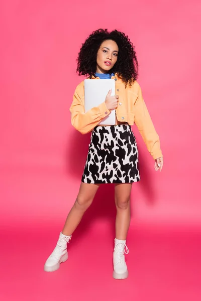 Volle Länge der lockigen Afroamerikanerin posiert mit Laptop auf rosa — Stockfoto