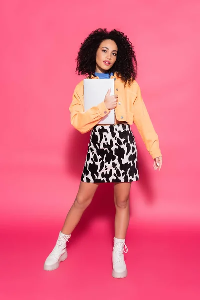 Longitud completa de rizado mujer afroamericana posando con el ordenador portátil en rosa - foto de stock