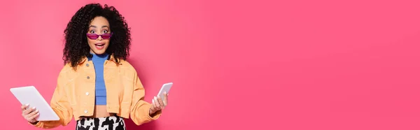 Femme afro-américaine confus dans des lunettes de soleil tenant smartphone et tablette numérique sur rose, bannière — Photo de stock