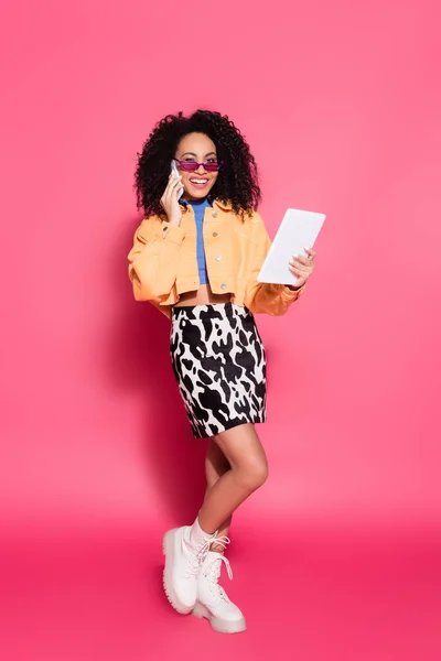 Longitud completa de la mujer afroamericana feliz en gafas de sol hablando en el teléfono inteligente mientras sostiene la tableta digital en rosa - foto de stock