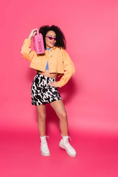 Longitud completa de la mujer afroamericana feliz en gafas de sol sosteniendo boombox y posando con la mano en la cadera en rosa — Stock Photo