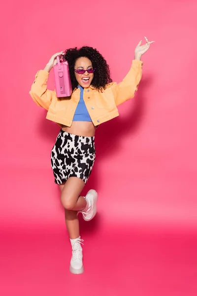Full length of happy african american woman in sunglasses holding boombox and posing on pink — Stock Photo