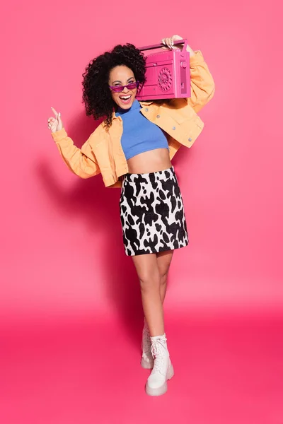 Full length of cheerful african american woman in sunglasses holding boombox and posing on pink — Stock Photo