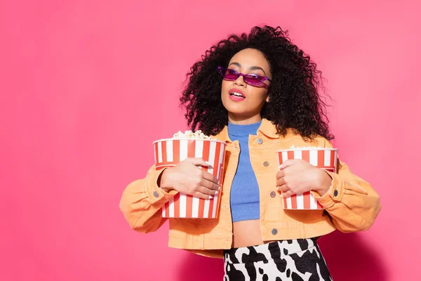 Afroamerikanerin mit Sonnenbrille hält Eimer mit Popcorn auf rosa — Stockfoto