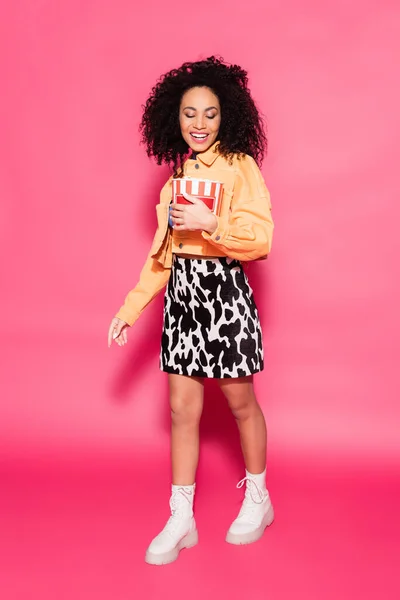 Longitud completa de alegre mujer afroamericana sosteniendo cubo con palomitas de maíz en rosa — Stock Photo