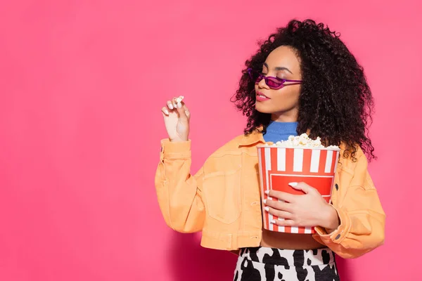 African american woman in sunglasses holding bucket and popcorn on pink — Stock Photo