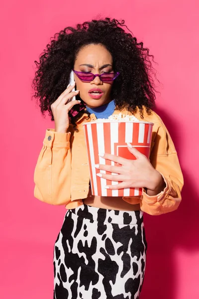 Afroamerikanerin mit Sonnenbrille hält Eimer Popcorn in der Hand und spricht auf Smartphone in rosa — Stockfoto