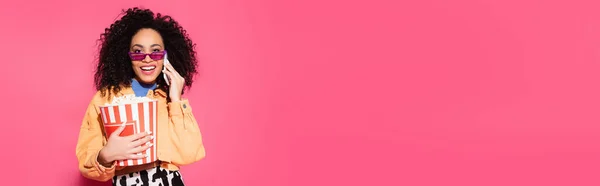 Щаслива афроамериканка в сонцезахисних окулярах тримає відро попкорну і розмовляє на смартфоні на рожевому, банер — стокове фото
