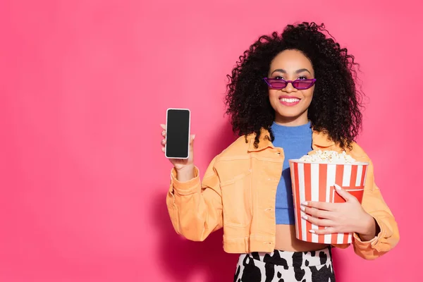 Smiling african american woman in sunglasses holding bucket of popcorn and smartphone with blank screen on pink — Stock Photo