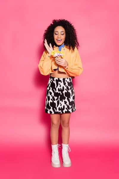 Pleine longueur de femme afro-américaine heureuse tenant un cône de gaufre avec des fleurs sur rose — Photo de stock