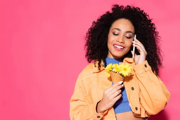 Happy african american woman talking on smartphone and holding waffle cone with flowers on pink — Stock Photo