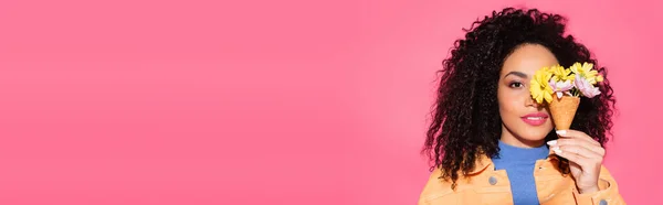 Кудрявая африканская американка закрывает глаза, держа в руках вафельный конус с цветами на розовом, баннер — стоковое фото