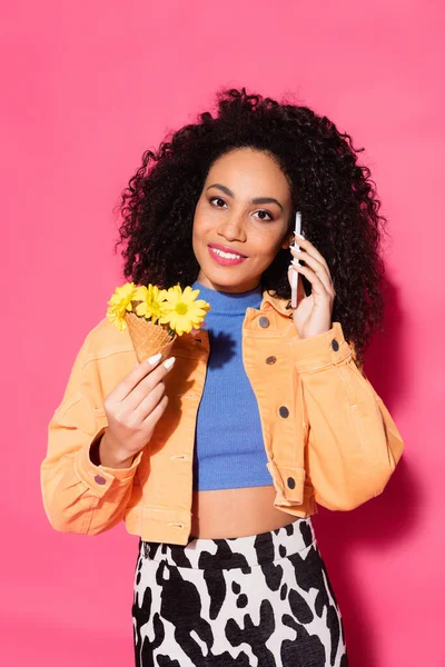 Happy african american woman talking on smartphone and holding waffle cone with flowers on pink — Stock Photo