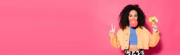 Curly african american woman blowing bubble gum, holding waffle cone with flowers and showing peace sign on pink, banner — Stock Photo