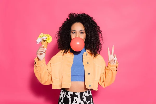 Curly african american woman blowing bubble gum, holding waffle cone with flowers and showing peace sign on pink — Stock Photo