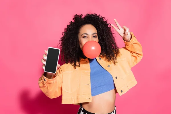 Mujer afroamericana rizada soplando goma de mascar, mostrando señal de paz y sosteniendo teléfono inteligente con pantalla en blanco en rosa - foto de stock