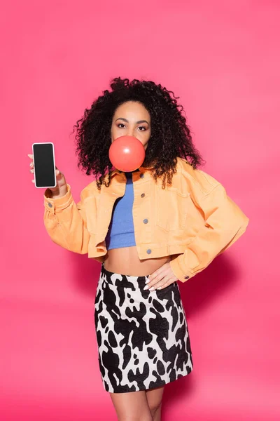 Mujer americana africana rizada soplando goma de mascar y sosteniendo teléfono inteligente con pantalla en blanco en rosa - foto de stock